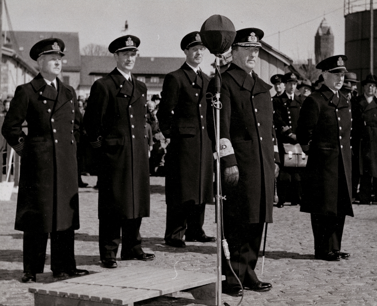 Fem uniformsklädda militärer av högre grad står framför en mikrofon. Kommendörkapten Lind af Hageby står som andra personen från vänster och konteramiral Samuelson som andra från höger. Bakom dem skymtar en folksamling.