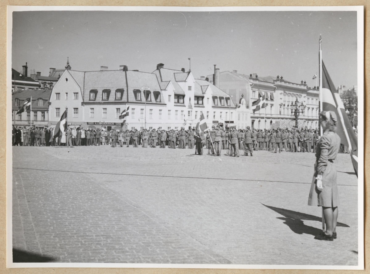 Lottor, soldater och hemvärnssoldater står uppställda på stortorget i Karlskrona. Till höger i förgrunden syns en lotta som håller i en svensk flagga. I bakgrunden syns byggnader runt stortorget, bland annat Karlskrona stadshotell.