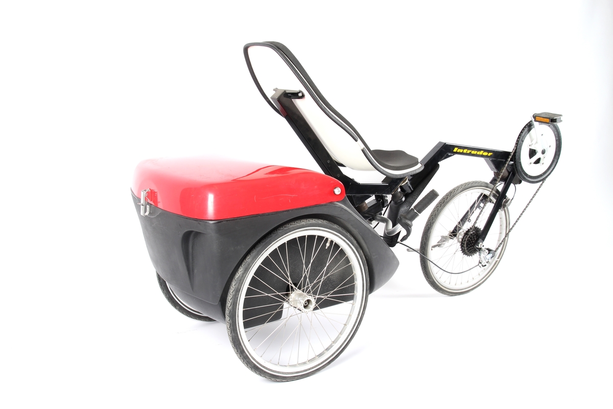 Rød og svart liggesykkel med 3 hjul og stor oppbevaringsboks bak. Sykkelen har felgbrems på forhjul og Sachs 7 girsystem.
