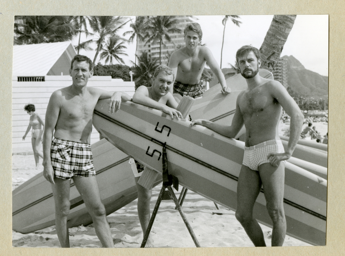 Bilden föreställer fyra män i badbyxor lutandes mot surfingbrädor på Waikiki beach. Längst till vänster står Oskar Linde i rutiga badbyxor. Bilden är tagen under minfartyget Älvsnabbens långresa 1966-1967.