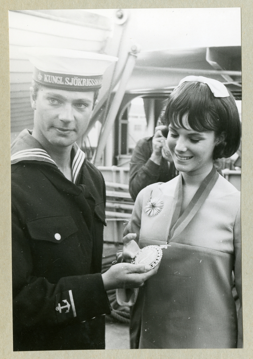 Bilden föreställer Carl XVI Gustaf i mörk uniform tillsammans med en kvinna i klänning. Runt halsen har kvinnan en medalj som Carl XVI Gustaf håller upp. Bilden är tagen under minfartyget Älvsnabbens långresa 1966-1967.