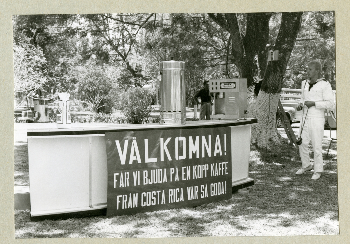 Bilden föreställer en besättningsman i vit uniform som står i ett grönområde där det serveras kaffe. På en skylt går det att läsa "VÄLKOMNA! FÅR VI BJUDA PÅ EN KOPP KAFFE FRÅN COSTA RICA VAR SÅ GODA!". Bilden är tagen under minfartyget Älvsnabbens långresa 1966-1967.