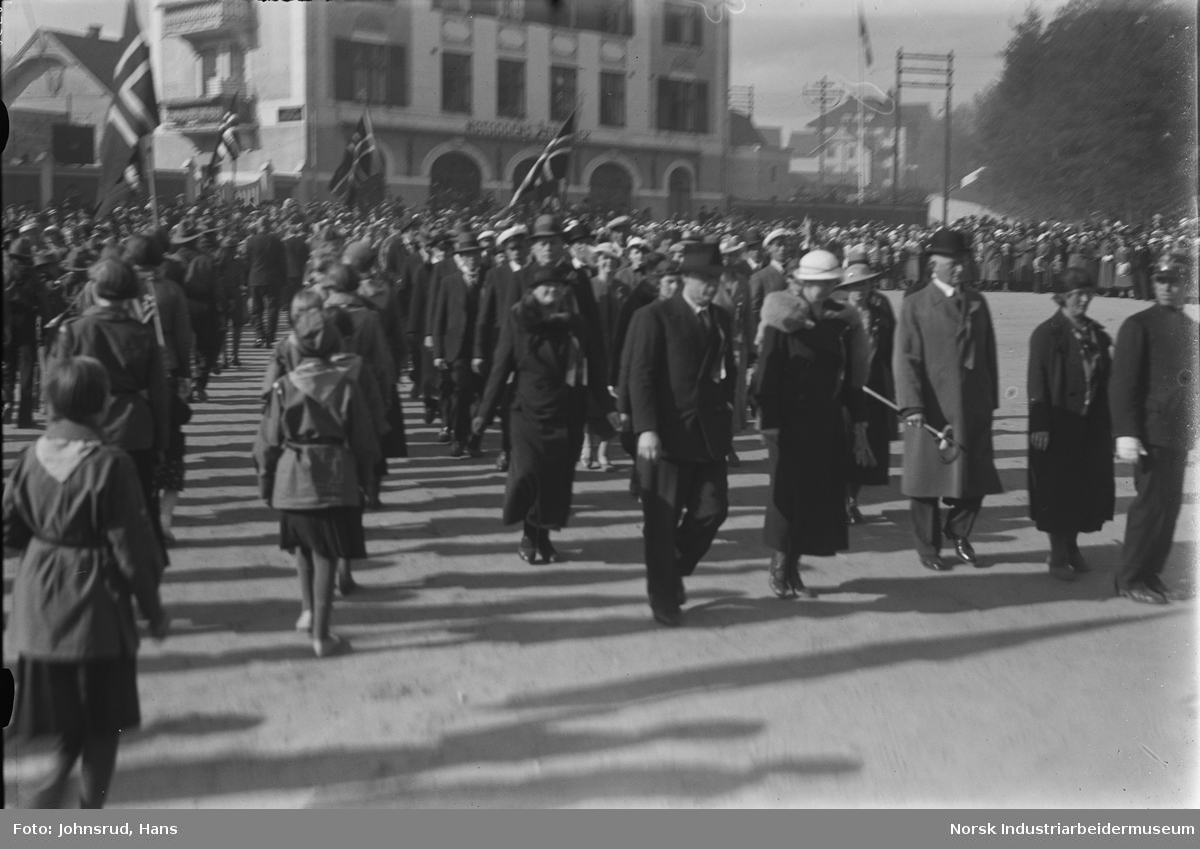 17. mai 1935. Barn og voksne i folketoget på torget. Voksne med hatt og 17. mai rosett. Folkemengde i bakgrunnen.