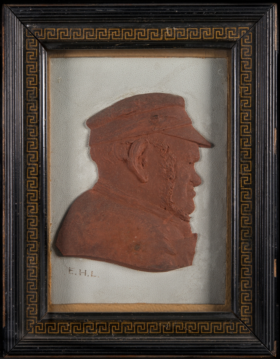 Relief i brun plastelin av "Gubben C. S. Ihrfors född 1832, död 1901", under glas, i svart ram.