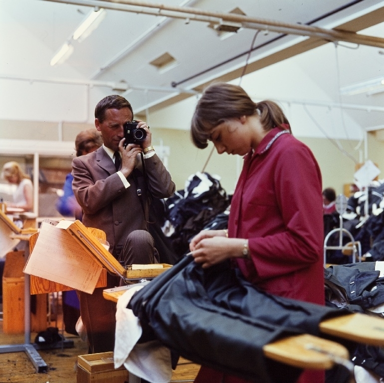 En syerske kontrollerer, klipper trådender og vrenger bukser i konfeksjonsfabrikken til Jonas Øglænd AS på Sandnes. Redaktøren i bedriftsavisen Thorvald S. Øglænd fotograferer arbeidet.