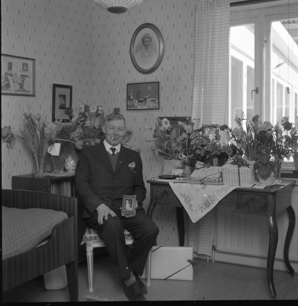 Louis Stackelberg fyller 50 år, mars 1964. Sitter på en stol omgiven av blomsterbuketter med en liten ramad tavla i händerna.