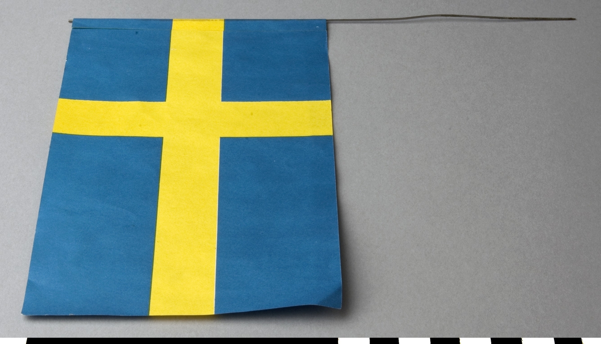 Flagga av papper, svensk. Flaggan är limmad på en ståltråd, som är avsedd att stickas fast i en gren. Trådens längd/höjd: c:a 220 mm.