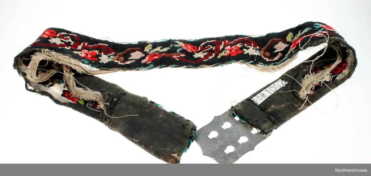Mannsbelte brodert på stramei med blomster- og bladmotiv i mange farger. Beltet er fôret med svart ullstoff og er påsydd ei sølvfarga metallspenne. Innleggsfor i lin. Beltet er svært slitt og fôret er revnet.