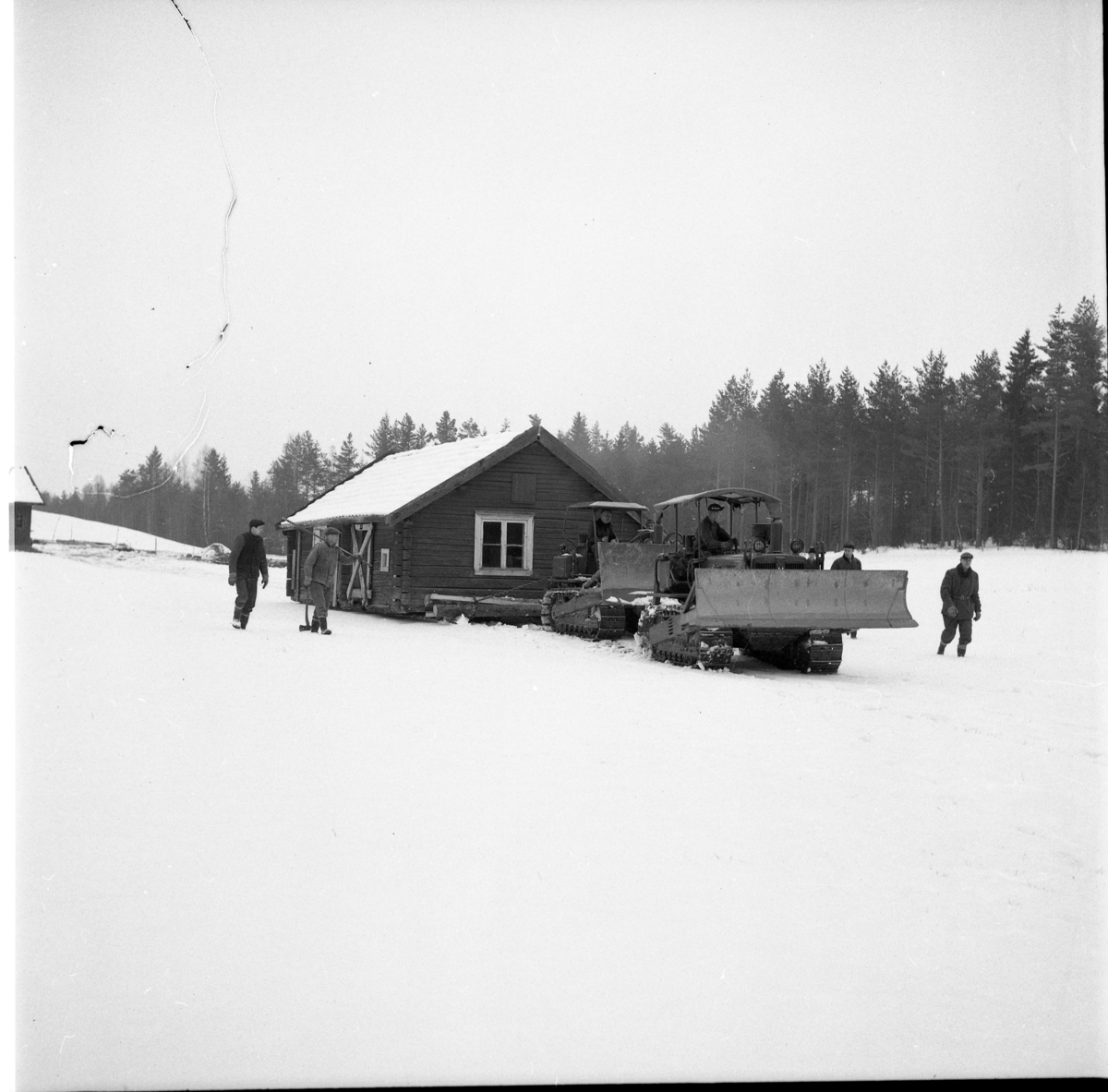 Husflyttning i Floddalen, Hullaryd. Två caterpillars i rad drar en timmerstuga över ett snöigt landskap, mars 1964.