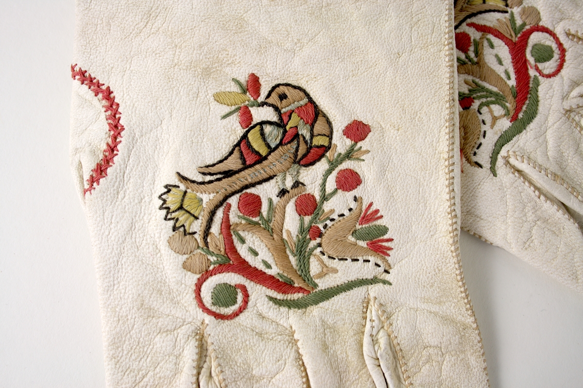 Handskar av vitt skinn med broderier som föreställer en fågel på en gren. 
Över tummens söm dekorerat med korsstygn i rött. Flikigt avslut mot handleden.