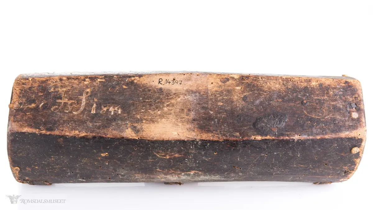 Uthulet trestokk som er høvlet flat i bunnen og har påsatte endestykker som er festet med trenagler. Tre lærremmer er spikret fast på tvers av åpningen. Rester av bek i bunnen.