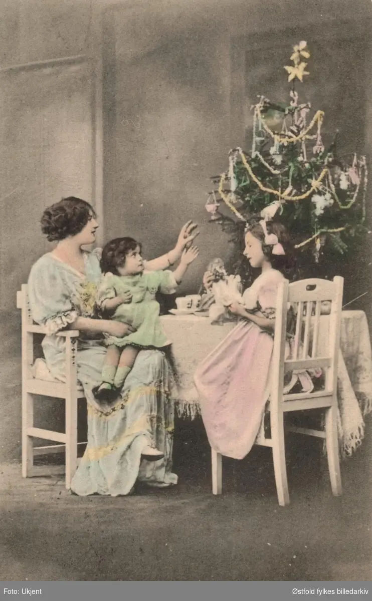 Julekort. Kolorert fotografi, tablå fra fotoatalier med juletre og mor med to barn. Poststempla 1913-14.