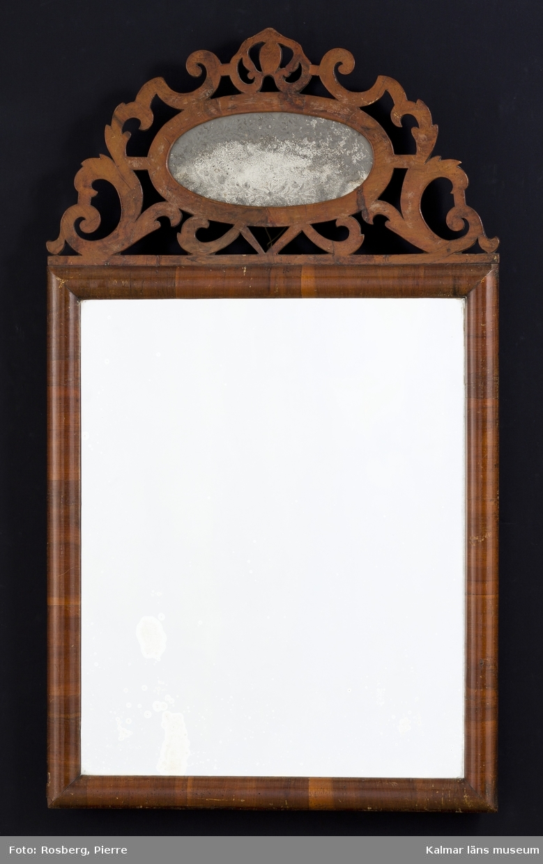 KLM 25153 Spegel av trä. Nytt glas, glas i överstycket ursprungligt, med etsad eller graverad baksida. Fyrkantig ram, halvrund list, fanerad i valnöt. Överstycke med genombruten rankdekor omfattande en mindre, oval spegel, vars glas har etsad eller graverad blomsterranka kring kanten. Datering: 1700-talets mitt eller förra del.