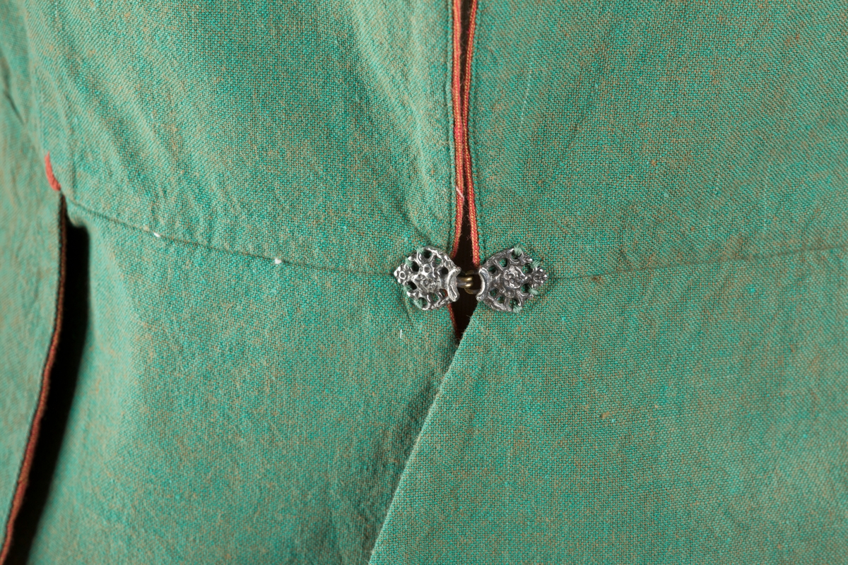 Grønt liv med rustrød kant og fór, seks skjøter. Håndsydd forsterkning i kontrastfarge øverst i skjøtesplittene. Lukkes med tinnhekte.