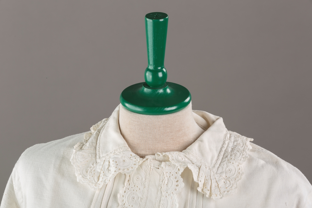 Form: Hoftelang trøye, Lange ermer, krage. Kragen og åpningen foran er kantet med  maskinprodusert blonde. Tre biser på hver side av åpn. foran. Lukkes med to løse knapper.