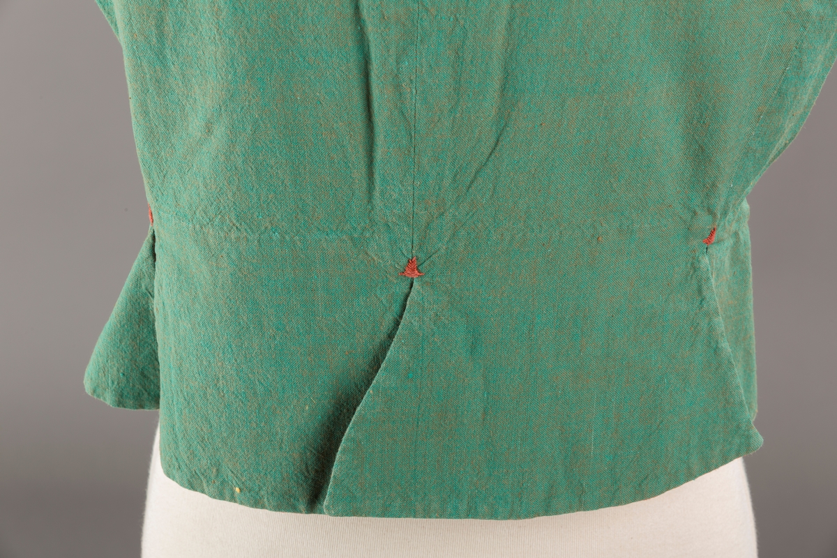 Grønt liv med rustrød kant og fór, seks skjøter. Håndsydd forsterkning i kontrastfarge øverst i skjøtesplittene. Lukkes med tinnhekte.