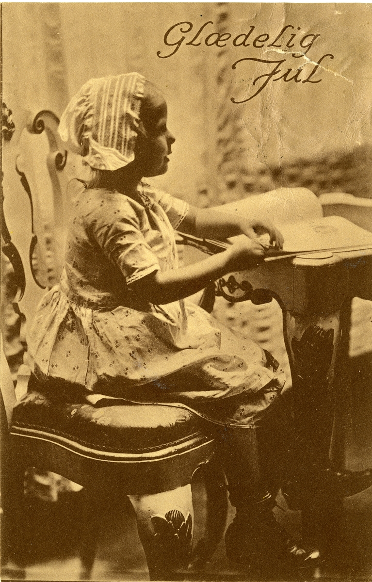 Julekort. Jule- og nyttårshilsen. Fotografisk motiv. Svart/hvitt. Ei jente sitter på en stol og leser i ei bok. Stemplet 24.12.1914.