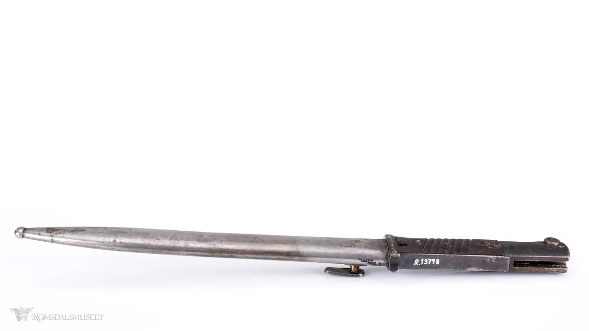 Metallhåndtak med lang pommel og geværfeste med fjæret knapp i bakkant. Serratert tregrep som er festet med 2 skruer. Langt spisst stålblad med egg og blodrand. Metallslire med kule enden og krok for festing til grunnutrustning/belte.