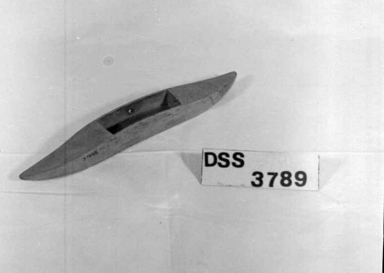 Avlang og buet vevskyttel av tre, med uthuling i midten der det er festet en trådspole av metall.