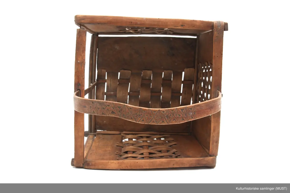 Firkantet trekurv med flettedekor og treskjæring på både kurv og håndtak. Dekoren består av to seksbladete blomster på hver side samt ulike trekantmønstre langs kanten av kurven og håndtaket. En av sidene i kurven mangler.