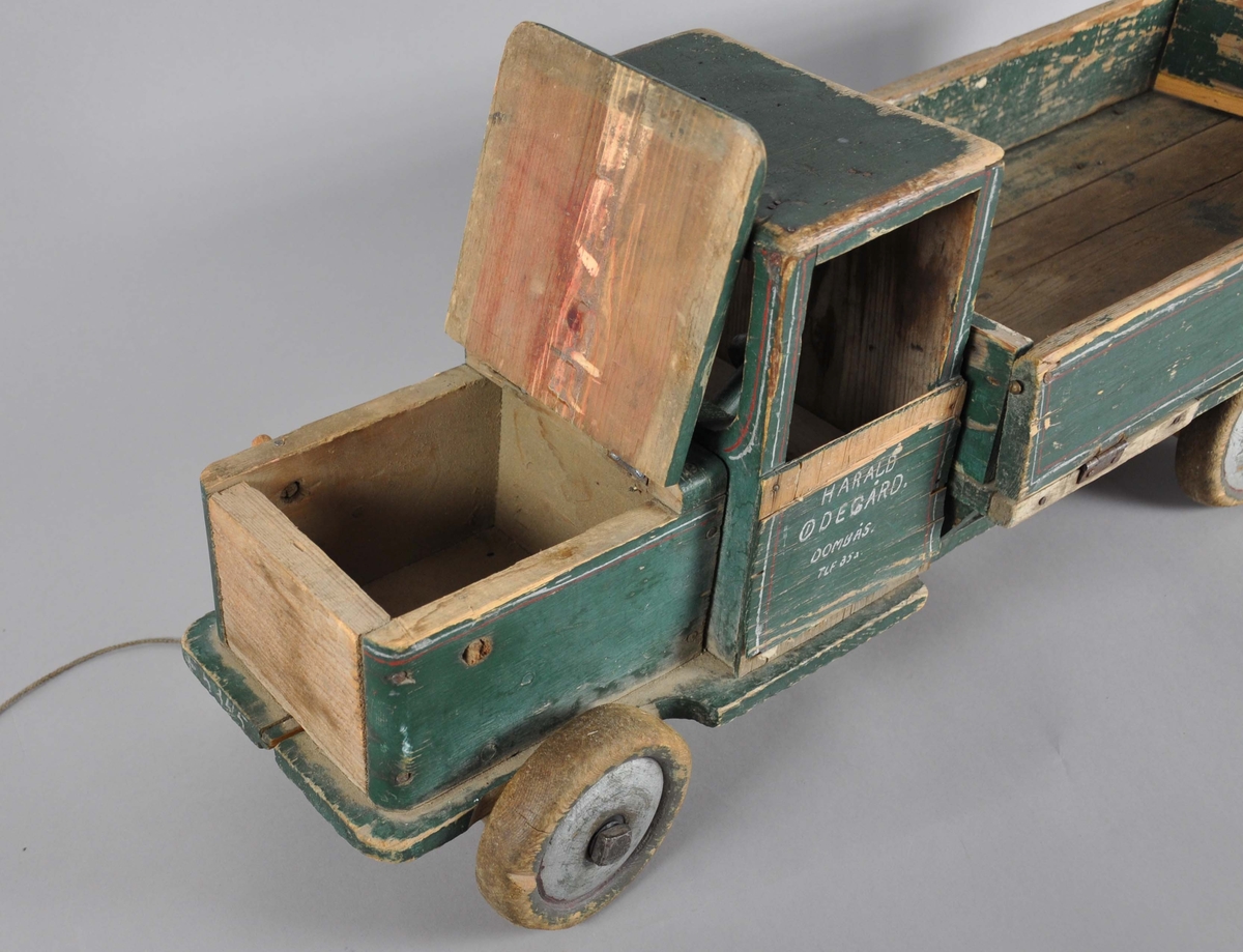 Håndlaget lastebil av tre. Består av frampart og lasteplan, samt bevegelige hjul. Lastebilen har en grønn grunnfarge med røde dekorstriper. Malingen er avslitt flere steder. Lasteplan og panser er hengslet slik at det kan tippes. Bak- og sidedør på lasteplanet er også hengslet slik at de kan legges ut. På framsiden står det en pinne ut på høyre side, på venstre side skal det være en tilsvarende pinne, men denne har knekt. På framsiden er det festet en tråd med pinne i enden, for å dra lastebilen.