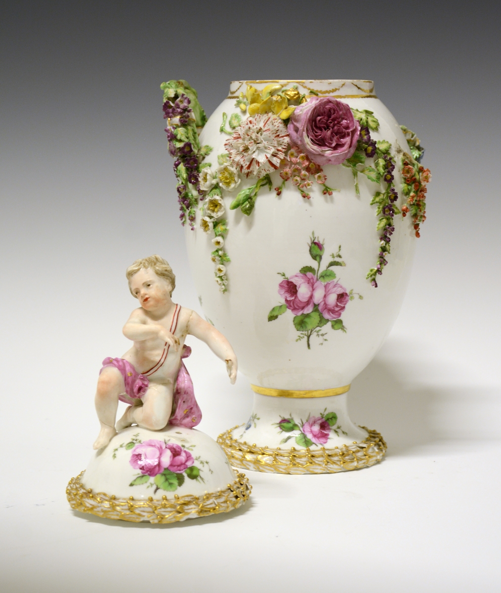 Potpourrikrukke av porselen med stett og lokk. Lokket er dekorert med håndmalt blomsterdekor og langs kanten en bord med blader i tre høyder dekorert med gull, over bladene gullfargede små kuler. På toppen av lokket en kjerub eller kupid som sitter på sitt ene kne. Han har på seg et rosa lendeklede og har holdt i et skjold eller en atributt. 
Urnens korpus er dekorert med håndmalte blomster, og modellerte blomster som sitter i klynger og strekker seg nedover korpus. De modellerte blomstene er uglasert og håndmalt. Urnen mangler en hanke. Den eksisterende hanken har modellerte blomster ved festene som strekker seg nedover i klynger.  Langs stettens kant samme bladbord som på lokket.