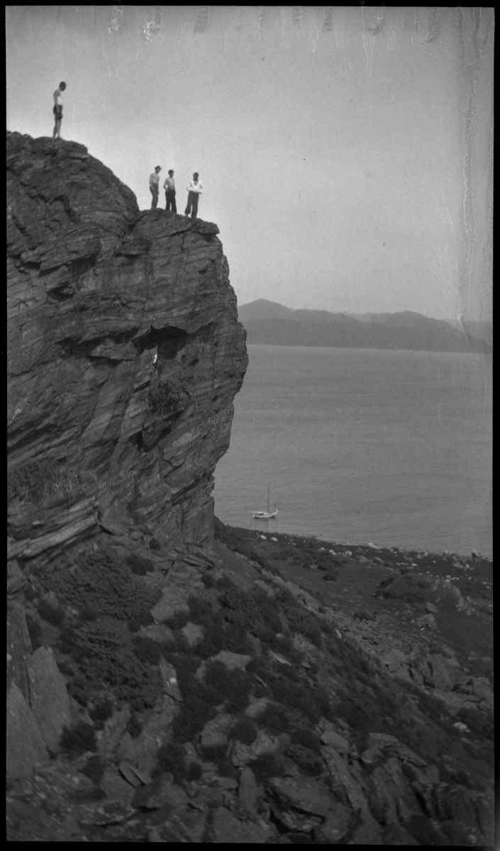 Harald Bergsaker, Ottar Roaldsøy, L. Fosse og Lindtner på den søndre enden av øya Hidle, øst for Stavanger. De er på kanten av et stup og hviler i gresset under stupet. En av dem ser utover sjøen med kikkert. Seilbåten "Vilja ligger i bakgrunnen.