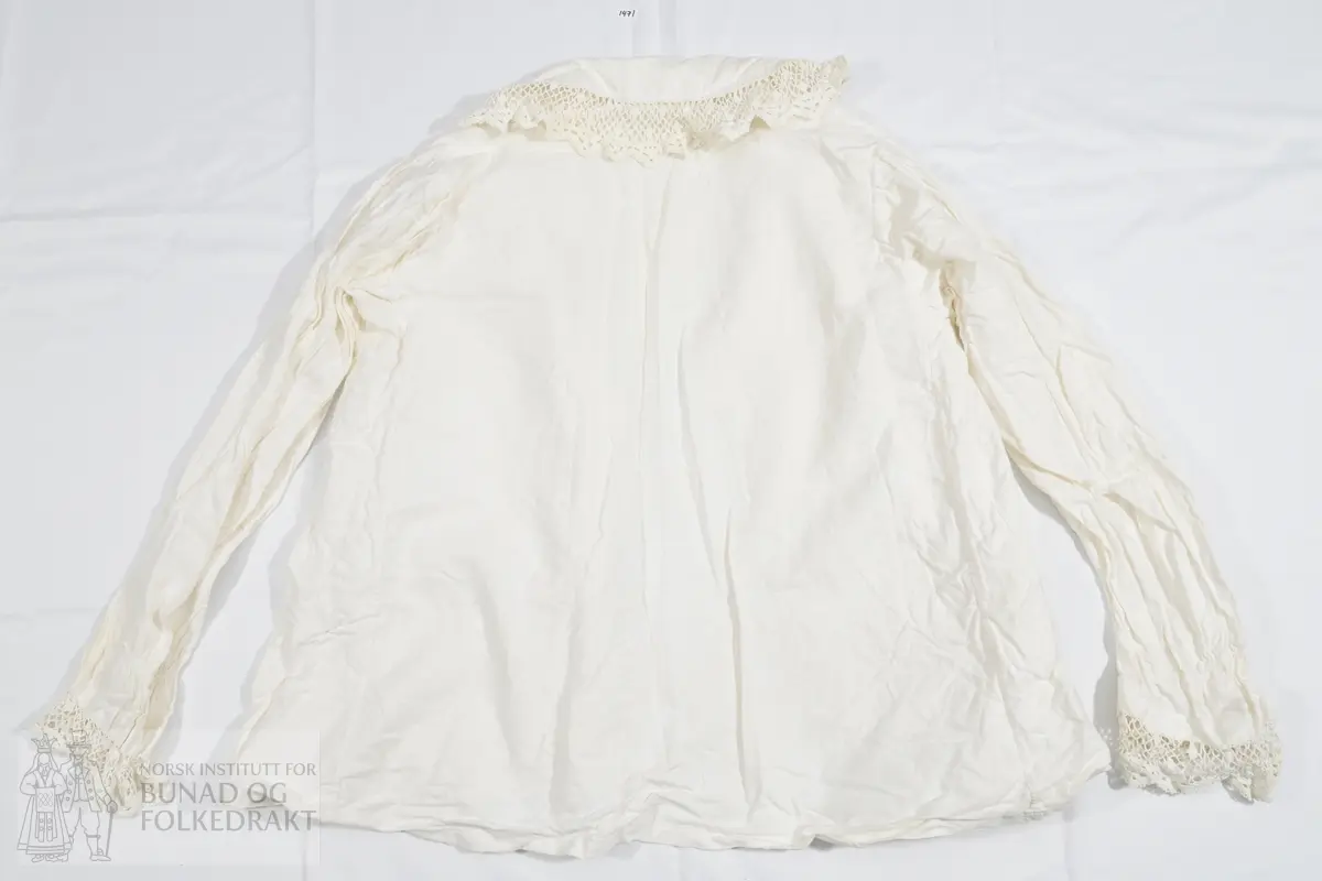 Skjorte i hvit bomullskypert med åpning midt fremme. Knapper trekt med bomullstoff og knapphull. Høyre knappestolpe har også påsydd en bred blonde. Halsen har bred linning, samt påsydd en bred krage med brede bomullsblonder. Blondene er heklet. Ensømserme. Ermet er rynket sammen ved håndleddet med et smalt bånd påsydd over rynkingen. Mansjett av samme, brede, blonden av bomull. Høyde heklet blondebestning: ca 4 cm.