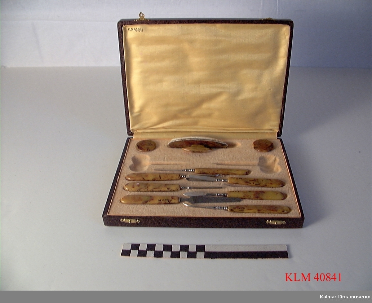 KLM 40841 Manikyrset, 10 delar, metall, konstmaterial. Redskapen har handtag av konstmaterial spräckligt i gult och brunt. Förvaras i tillhörande etui med fördjupningar för varje redskap. 2 saxar saknas.