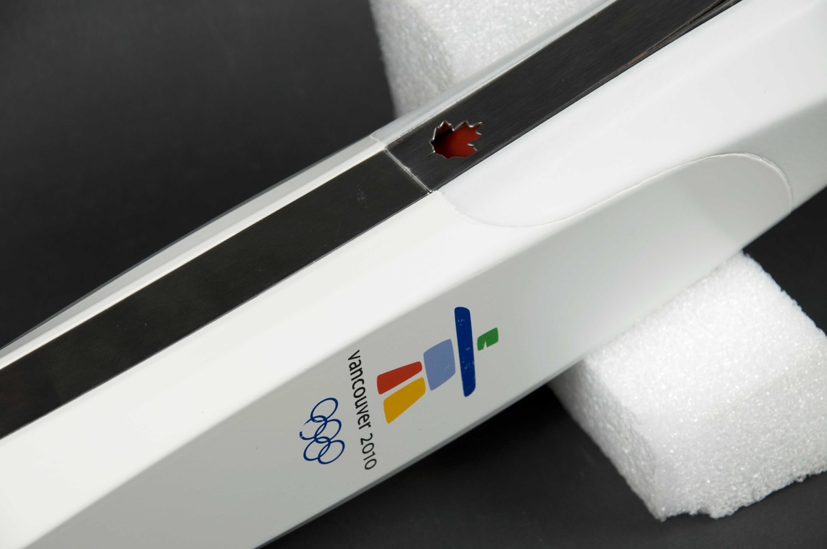 Hvit håndfakkel av plast og metall. Fakkelen har emblemet for de olympiske vinterleker i Vancouver i 2010. Det er også motiv av lønneblad på fakkelen.