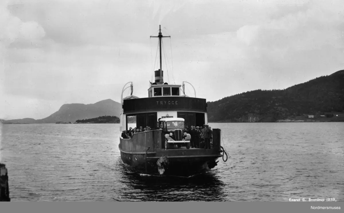 Bilferga Trygge med bil T-2286. Grillen tilsier at dette trolig er en Ford V8 1937-modell lastebil, forøvrig med dobbelt førerhus. Det er trolig Aarø Auto i Molde som eier lastebilen. Det er forøvrig mest passasjerer ombord. Postkort Georg Sverdrup 1939. Kristiansund. Gjemnes-ruta? - B/F "Trygge" ble bygd ved Storvik Mek. Verksted i Kristiansund og levert i 1938. Den var bygd for ruta Kristiansund-Kvernes-Gjemnes-Torvikbukt som dermed kunne avløse tidligere båtrute på strekningen. "Trygge" gikk i denne ruta til 1949 da den ble avløst av B/F "Gjemnes". 
T-2286 var et registreringsnummer som ble brukt av Aarø Automobilselskap i Molde. I 1934 ble nummeret tatt i bruk på en Ford postbil. Aarø Auto hadde kjørt post Molde-Batnfjord fra 1908, og fra 1932 Molde-Gjemnes. Det var omlasting til og fra båt videre til Kristiansund og Trondheim. I 1938 kunne postbilen følge ferje til og fra Kristiansund. Det samme gjorde gods- og melkerutene til og fra Batnfjorden som Batnfjord Auto drev. Personbussene gikk bare til og fra Gjemnes enda. Først i 1952 ble ferjestrekningen forkortet til Kvitnes-Gjemnes og personbussene kunne kjøre passasjerene hele veien Kristiansund-Molde. (Info fra Sveinung Berild 2018.)  Nordmøre museums fotosamling.