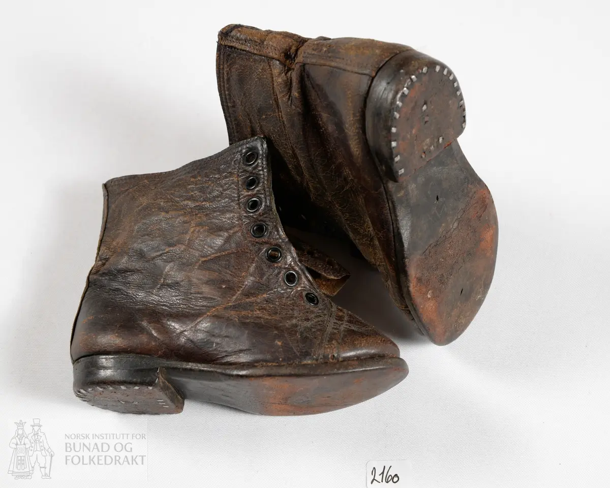 Barnesko av brunt skinn. Lav støvelmodell. Løs tunge som har vært festet til skoene med lissene, lissene mangler nå. 7 maljer på hver side, til sammen 14 stykk, til lissene. Tungene har takkekant øverst. Foret med et stripete bomullsstoff i i kypertbinding, fargene er beige og mørk blå. Sålene er av lær. All synlig søm er gjort på maskin.