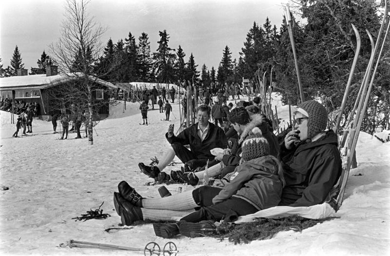 Svart-hvitt foto av vinterkledde mennesker som har satt skiene i snøen og tar seg en rast i påskesola ved Lageråkvisla skihytte i Vang almenning.