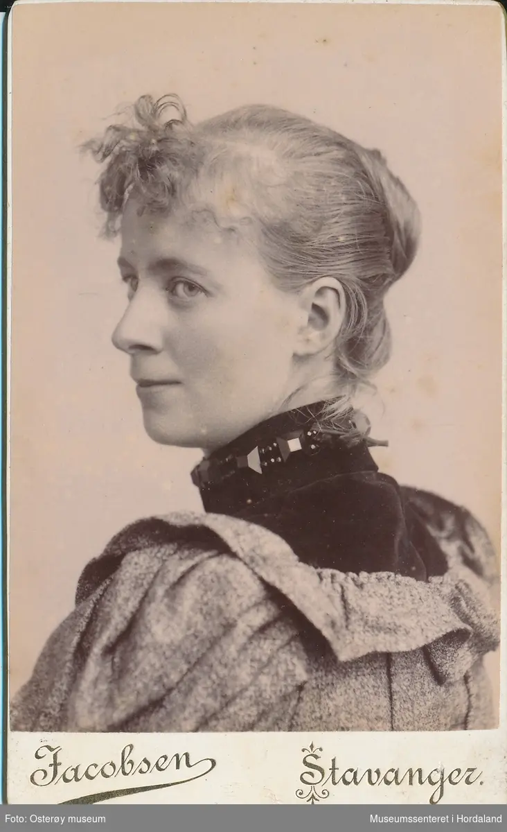 portrettfotografi av ung kvinne i profil med håret rulla opp i nakken og krølla lugg, kledd i mønstra bluse med svart høghalsa genser inni