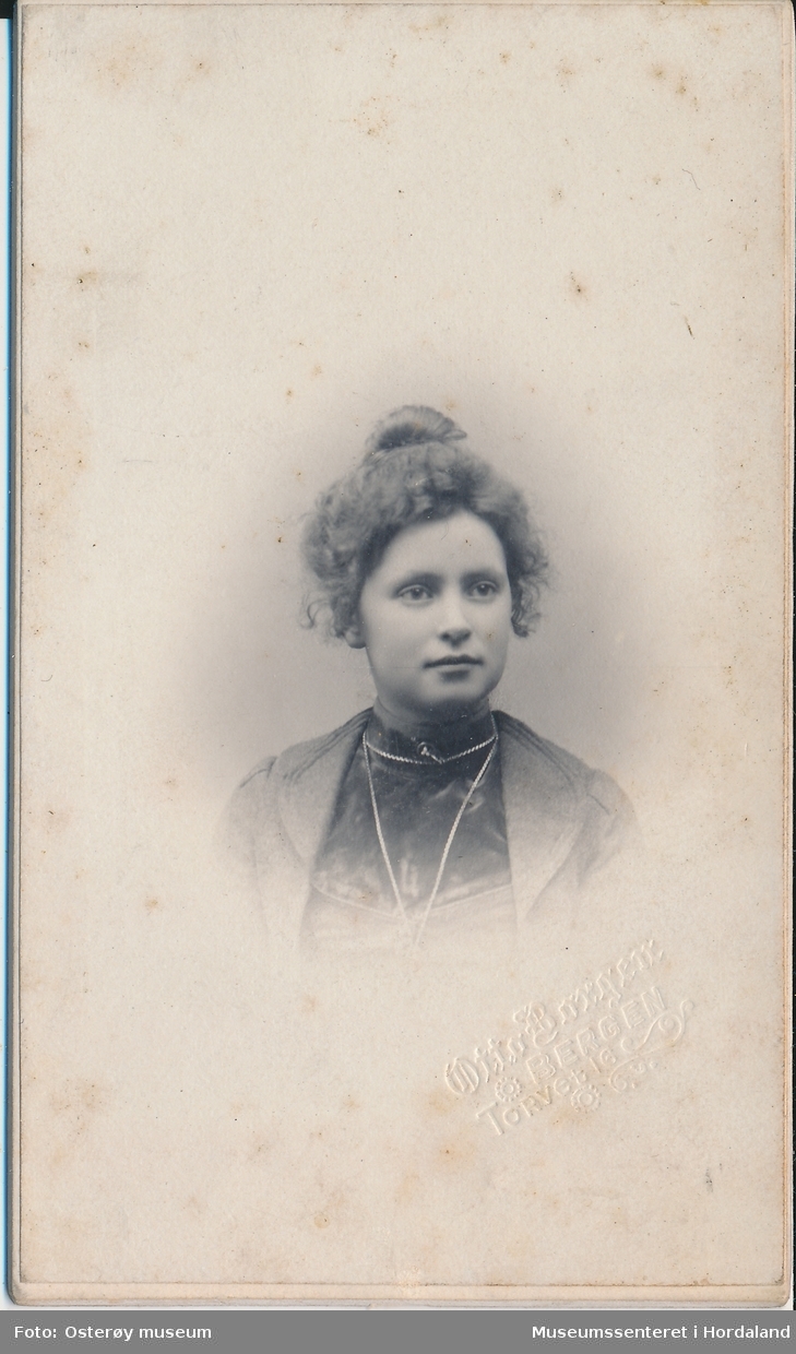 portrettfotografi av ung kvinne med krølla hår rulla opp på hove, ljos jakke og mørkare bluse med kjede om halsen