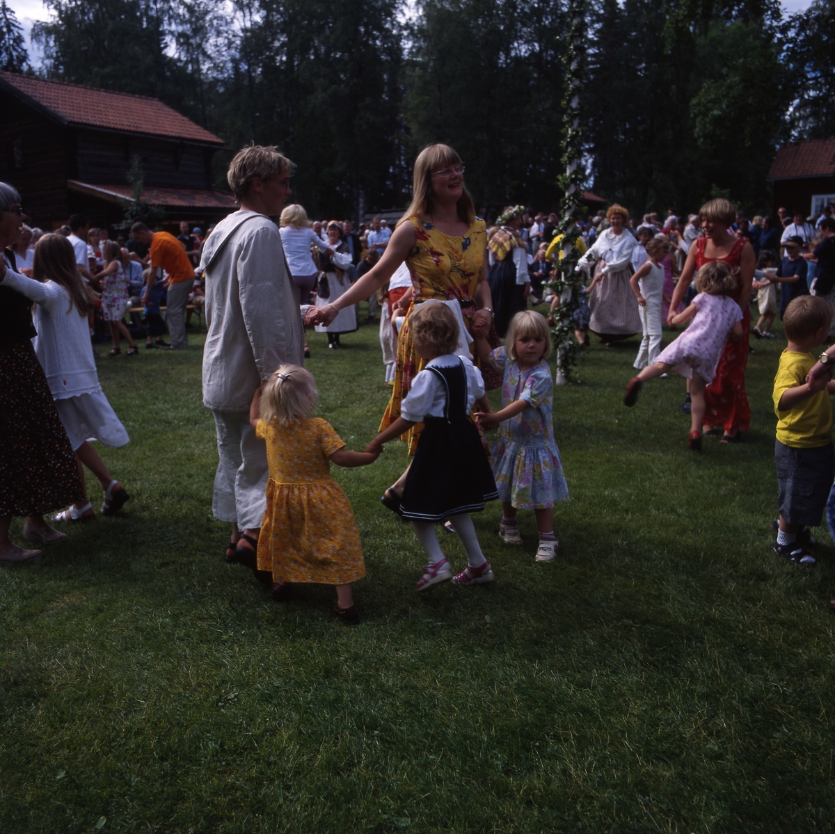 Midsommarfest vid Västerby hembygdsby i Rengsjö år 2000. Solen lyser och några vuxna och barn dansar i en liten ring vid midsommarstången.