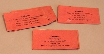 Sällskapsspel bestående av frågekort. Frågekorten är av ljusrött papper med svart tryck.