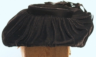 Damhatt sydd av svart sammet.Sydd i s.k. barettmodell utan skärm.Tyget lagt i mjuka veck. Prydd i nacken med en rosett av sammet samt svarta och rosa fjädrar.