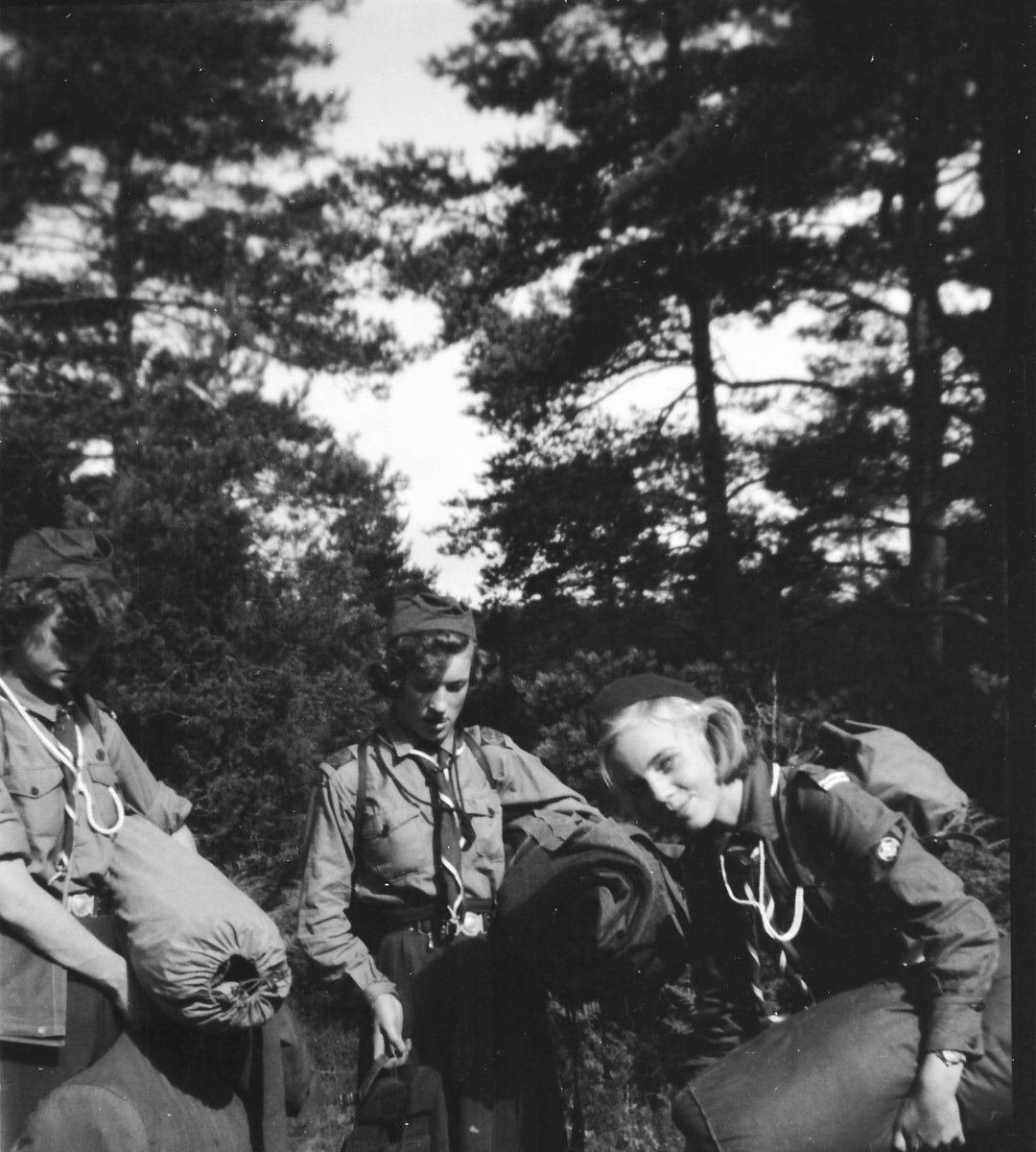 Tre scoutflickor i skogen med tält och annan packning.