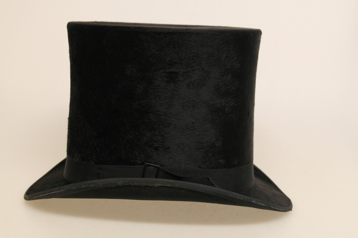 Svart flosshatt i en type fløyel (kastor/beverhår?). Hatten har svart ripsbånd med svart, rektangulær spenne festet på den ene siden. Innvendig er hatten kledd med papir og et silkeaktig stoff i purpur. I bunnen er det et gull-/kobberfarget stempel.