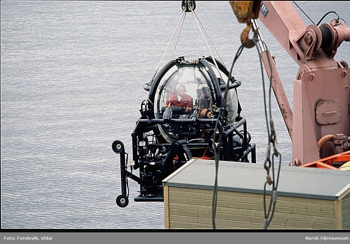 Miniubåten Check Mate til NUTEC holdes over vannoverflaten med en kran på kaien til NUI's dykkesenter i Gravdal. To personer bemanner ubåten.