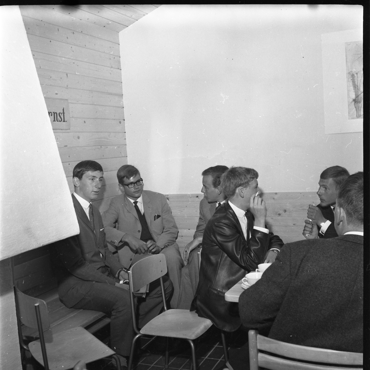 Grännaskolans första elever i skolans gillestuga i samband med avslutning 1964. Till vänster sitter Anders Hugander intill Jan Lothigius med glasögon. Andra elever runt ett bord med kaffekoppar.