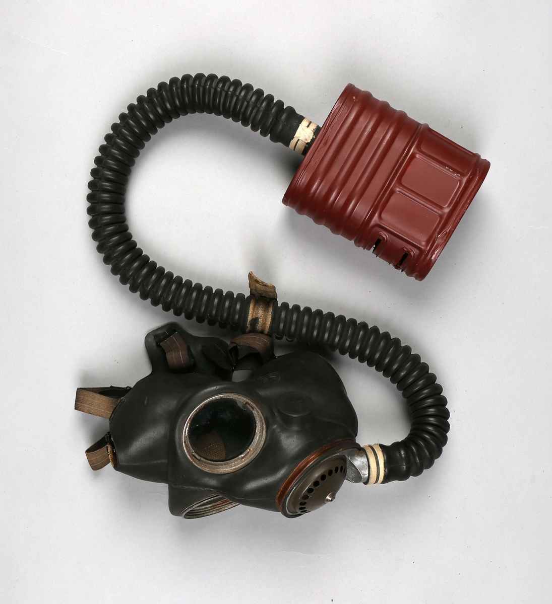 Gassmaske i tøyveske. Gassmaske i gummi med beholder av metall som er koblet sammen med en fleksibel slange. Oppbevares i tøyveske.