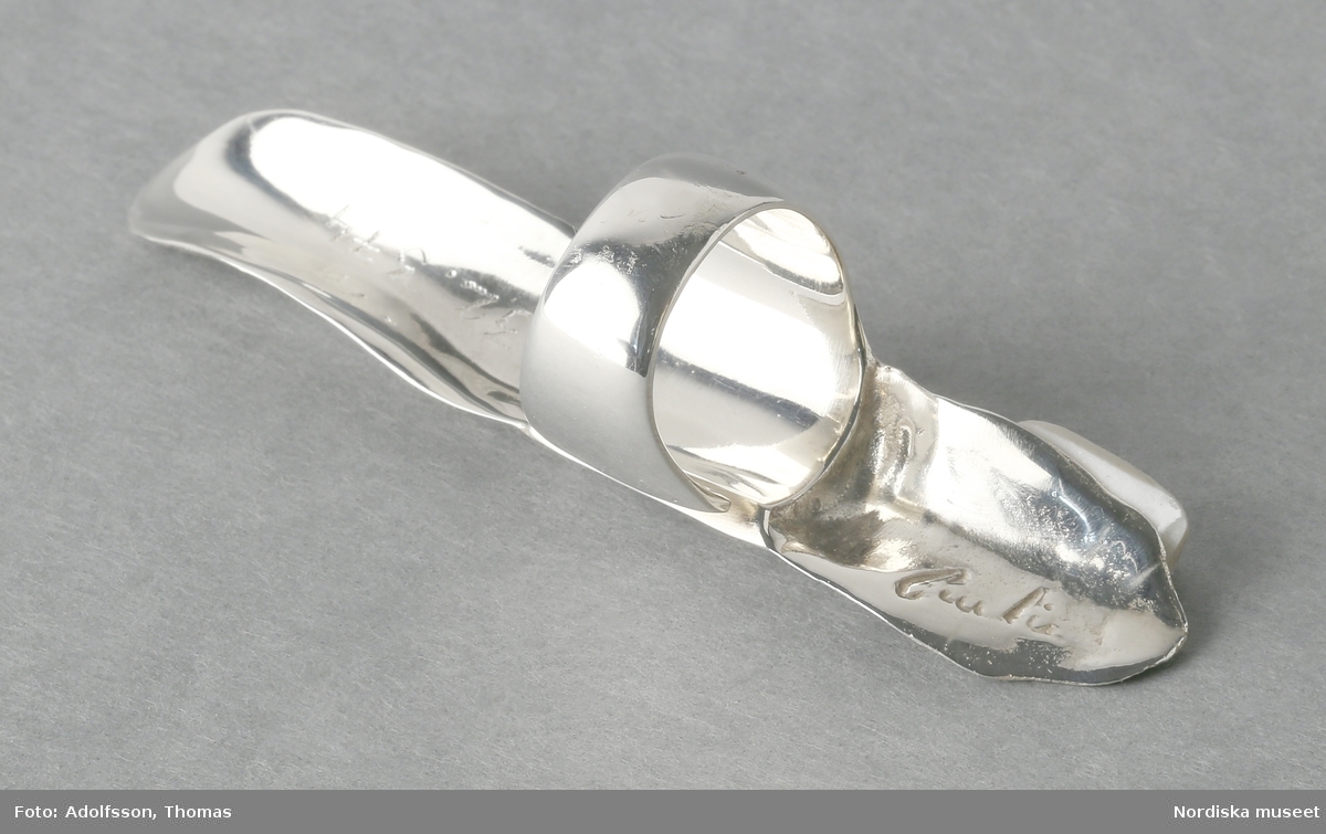 Ring av silver, överdel i form av avlång avgjutning av fingerrygg. Underdel bestående bred ring med slits på ena sidan. Undertill graverad "Cornelia".
/Leif Wallin 2018-02-09