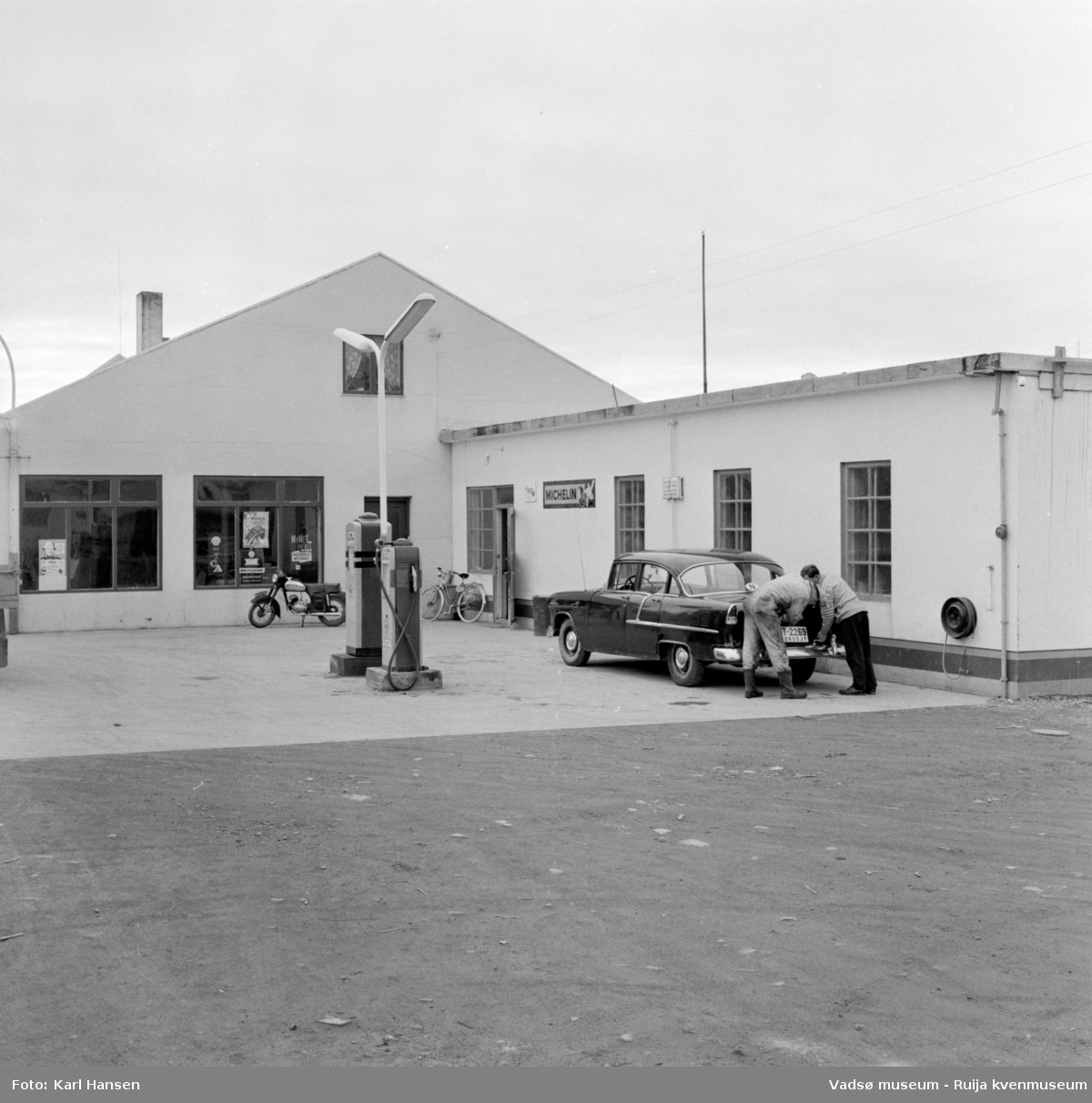 Vadsø sentrum 1959. Shell Bensinstasjon og tankanlegg i Tollbugata. Reklameskilt, moped, drivstoffpumper og en drosje i forkant av bygningen.