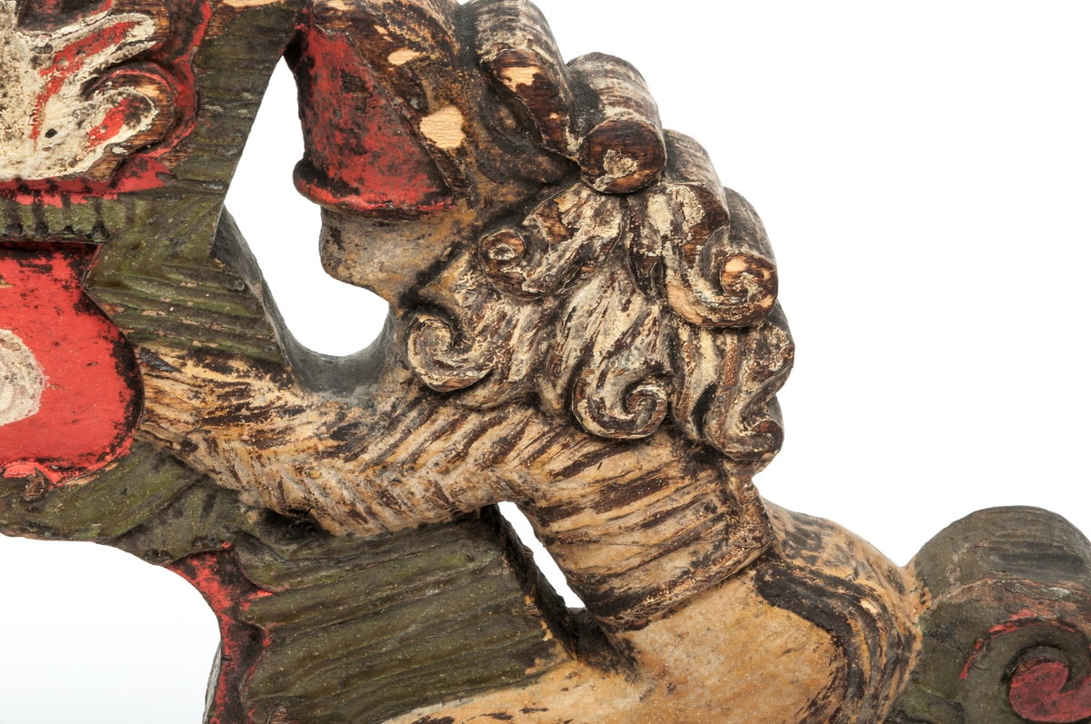 Selkrok av trä, skulpterad och målad i rött, grönt och brunt. Skulpteringen föreställer två lejon hållande ett kronliknande ornament.