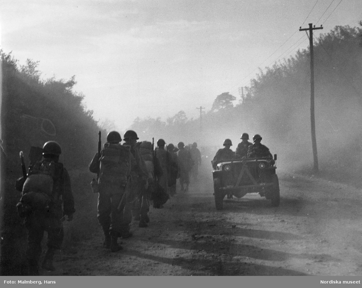 Koreakriget. En kolonn med amerikanska soldater marscherar på en dammig väg, en jeep passerar i motsatt riktning.