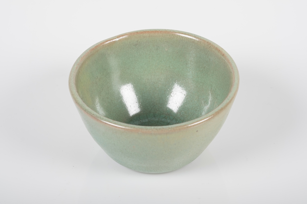 Rund kopp i keramikk med grønn lasur. Buet hank på koppen. Tre små knotter på undersiden av koppen, usikker funksjon.