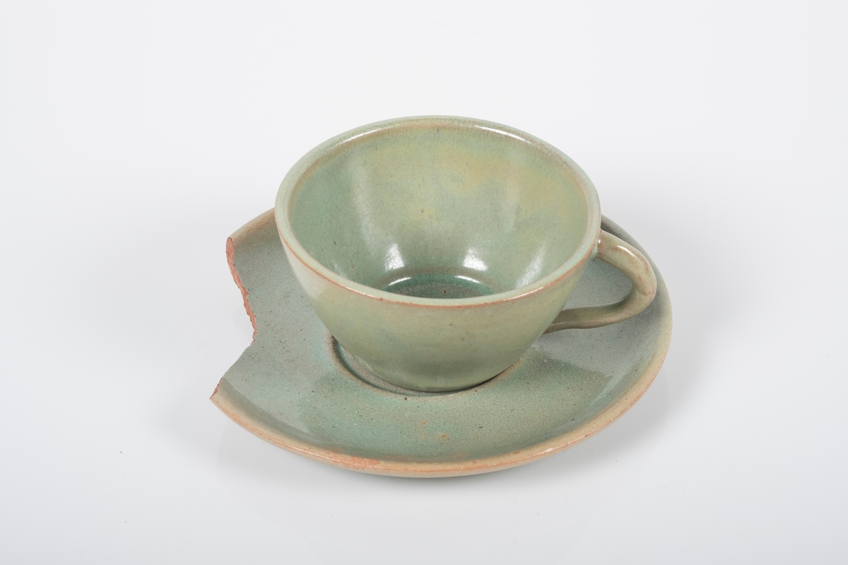 Kopp med skål i keramikk med grønn lasur. Buet hank på koppen. Koppen og skålen har spor etter tre knotter på bunnen, usikker funksjon. Bunnen på skålen har matt overflate. En brukket del av skålen mangler.