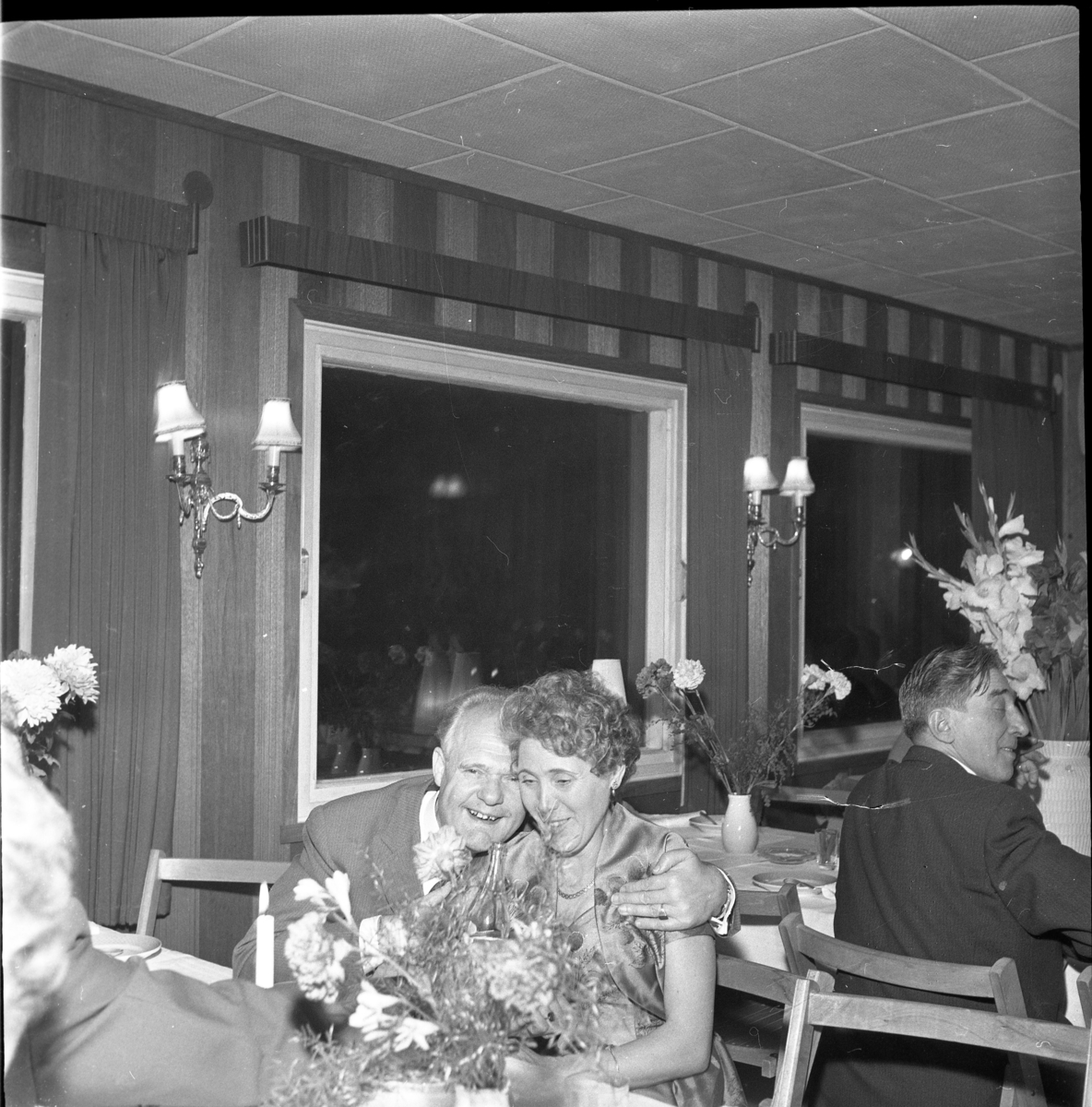 Sittande bord med blommor och tända ljus. Holger Andersson kramar om Birgit Heldebrandt. Vid bordet bakom sitter Rudolf Johannesson. Man firar troligen Gerhard Palms 50-årsdag.